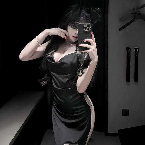 Woman in black satin qqny pure & sexy knotted neckline sleep dress, body-con fit, posing with phone in dim room.