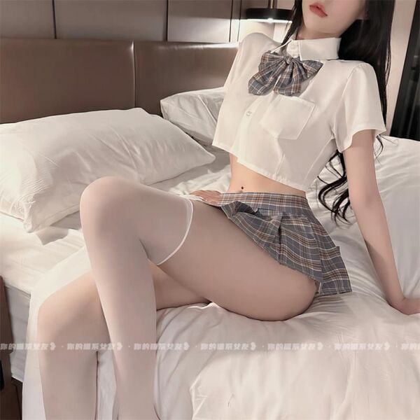 Woman wearing a plus size sexy jk uniform set with a white crop top, plaid bow tie, and seductive pleated mini skirt on a bed...