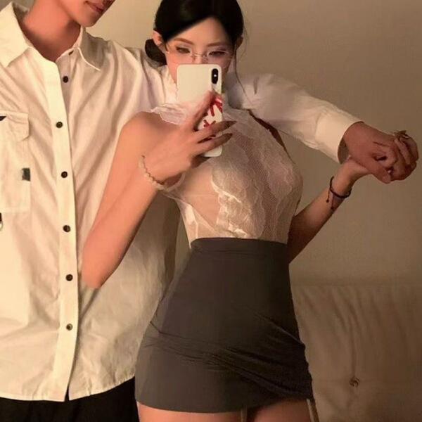 Woman in white lace bodycon dress with gray skirt, posing seductively while taking a mirror selfie with partner nearby.