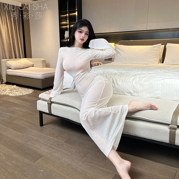 A woman poses elegantly on a bed wearing a sheer white long-sleeve nightgown dress, highlighting its form-fitting design and ...