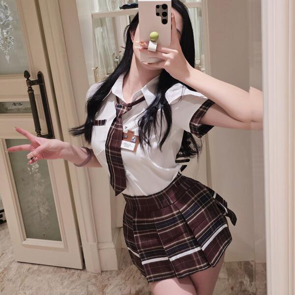 A woman poses in a mirror wearing a cosplay student uniform outfit, featuring a white blouse with a tie and a plaid skirt, em...