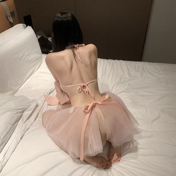 Woman wearing pink cherry romance lingerie set with bow details, sitting on bed, back view, sheer tulle skirt.