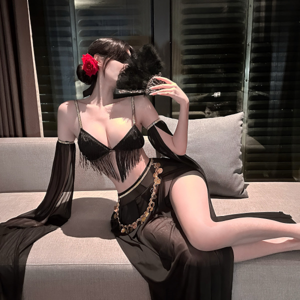 Woman in black fringe lingerie set with sheer sleeves, gold chain belt, and red rose hair accessory on sofa.