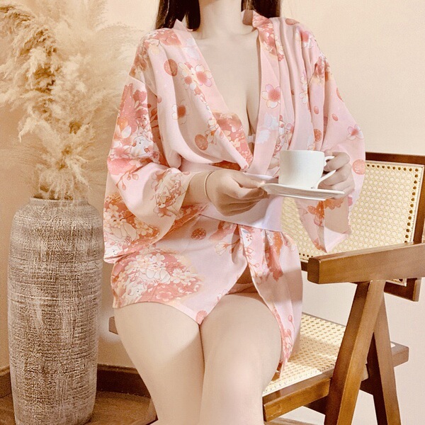 A woman sits on a chair holding a cup, wearing a pink floral japanese kimono-style robe. the outfit features a deep v-necklin...