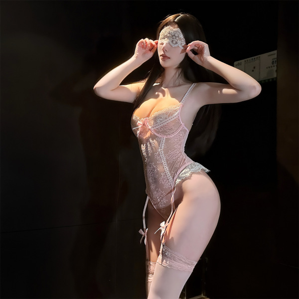 Woman wearing pink luxury lace sensual bodysuit with sheer panels, lace trim, and matching eye mask in dim lighting.