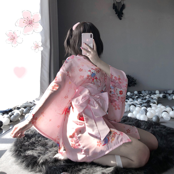 A woman sits on a fur rug wearing a pink floral japanese-style kimono robe set, featuring lace details and a bow at the waist...