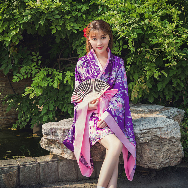 A woman wearing a purple lolita dress with cherry blossom patterns, paired with a pink ribbon and fan, sitting by a pond surr...