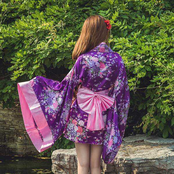 A woman stands by a pond wearing a purple lolita dress with cherry blossom patterns, featuring a pink ribbon belt and flowing...