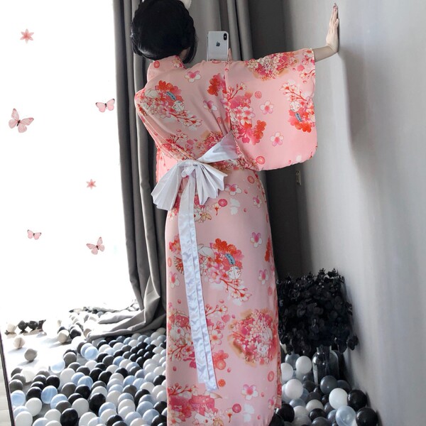 A woman stands by a window wearing a pink floral japanese kimono-style dress with a white ribbon belt, set against a backdrop...