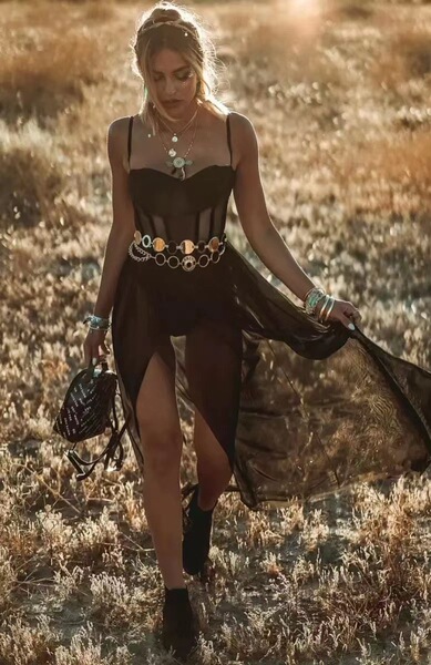A woman walks through a field wearing a black muse maxi dress with white mesh details, featuring a high slit and flowing fabr...
