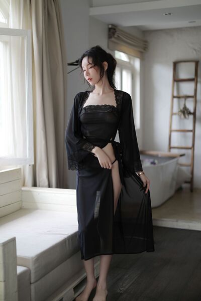 A woman stands by a window wearing a black lace sleep set, featuring a sheer nightgown and matching bralette, perfect for sed...