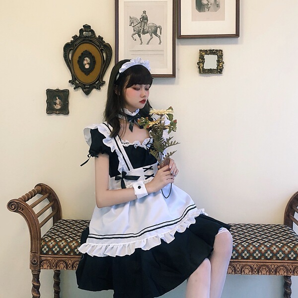 A person wearing a black and white lolita maid dress with ruffles, sitting on a bench holding flowers against a vintage wall ...