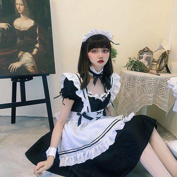A woman wearing a black and white lolita maid dress with ruffles, sitting indoors next to an easel painting. the outfit featu...