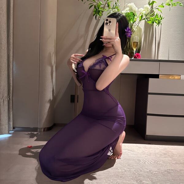 Woman in purple sheer nightsigh nightseduce lingerie set, kneeling by vanity with flowers and champagne glass.