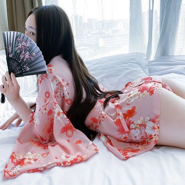 A woman lounges on a bed wearing a pink plus-size japanese kimono with floral prints, featuring a deep v neckline and flowing...