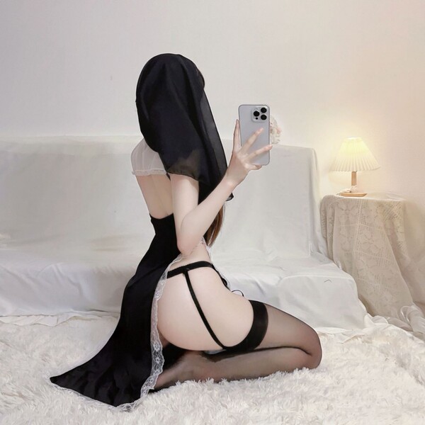 Woman in black plus size nun lingerie with cut-outs and high slit, posing on bed for sexy role play at home.