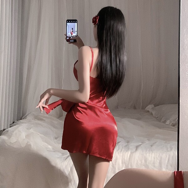Woman in red satin chemise nightgown with adjustable straps, posing by bed, holding phone for mirror selfie.