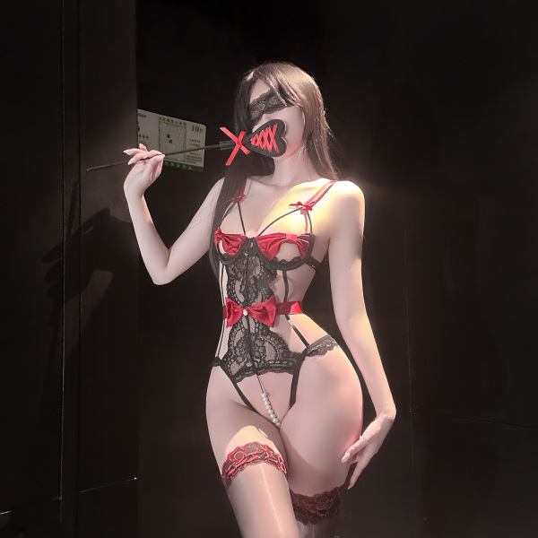 Woman wearing red and black lace lingerie set with bows and pearl bodysuit, holding a wand in a dimly lit room.