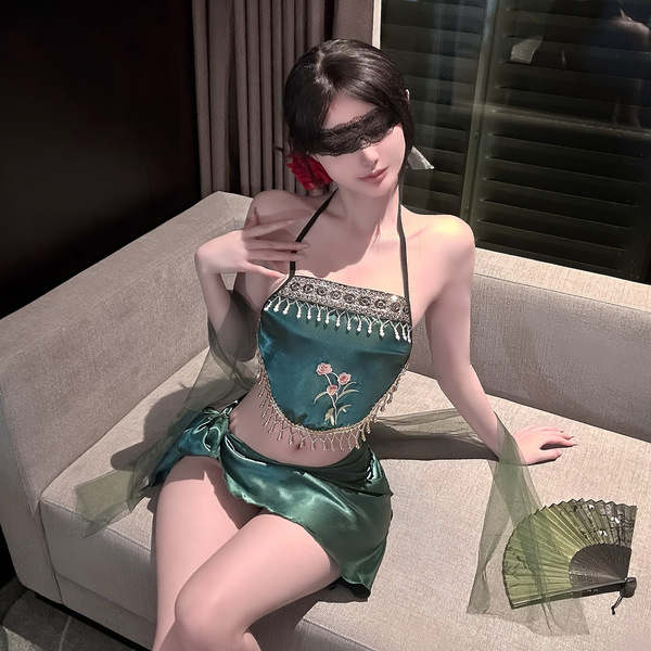 A woman poses on a couch wearing a green, exotic-style bralette with floral embroidery and fringe details. the outfit feature...