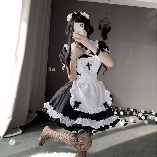 A person wearing a black and white maid costume set, featuring ruffled apron, fishnet stockings, and headband, posing indoors...