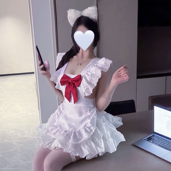A person wearing a white maid costume with a red bow, fluffy cat ears, and ruffled details sits next to an open laptop. the o...