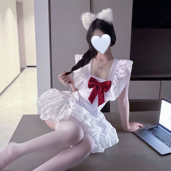 A person wearing a white cosplay maid costume with a red bow, fluffy ears, and ruffled details sits beside an open laptop.