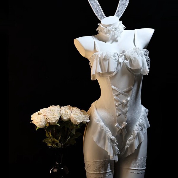 White bunny girl cosplay lingerie set with lace, ruffles, and open-crotch design on mannequin beside white roses.