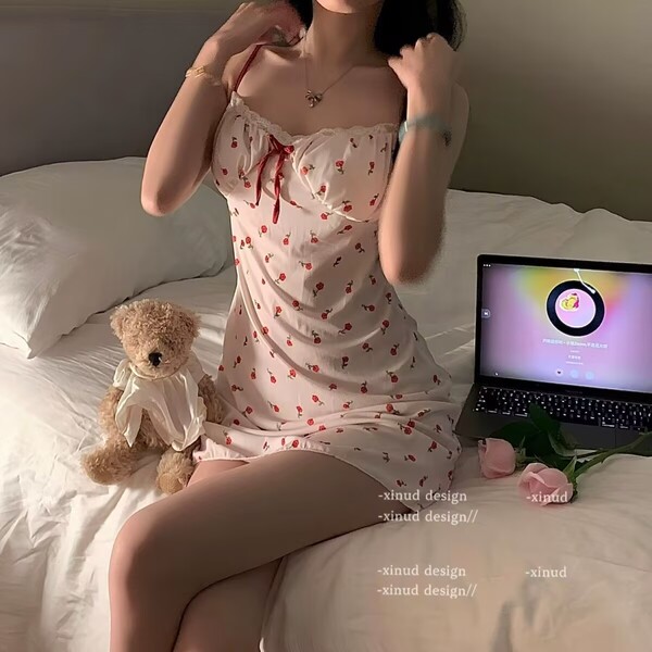 A woman sits on a bed wearing a cherry print cami slip dress with lace trim and built-in pads, accompanied by a teddy bear an...