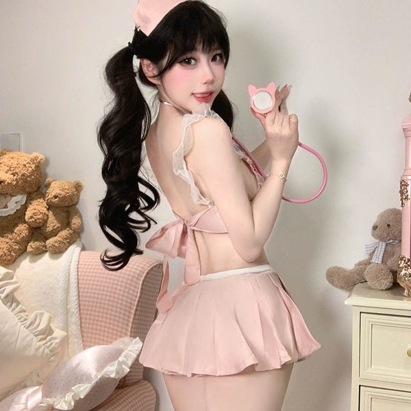 Woman in pink seductive nurse costume with bow back, holding stethoscope, surrounded by teddy bears and soft decor.