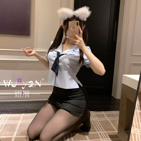 Woman kneeling in student-themed lingerie set with white blouse, black skirt, sheer tights, and fluffy cat ears headband.