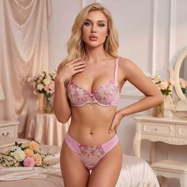 Woman modeling pink sheer lace lingerie set with floral embroidery, featuring a bra and matching panties in a romantic bedroo...