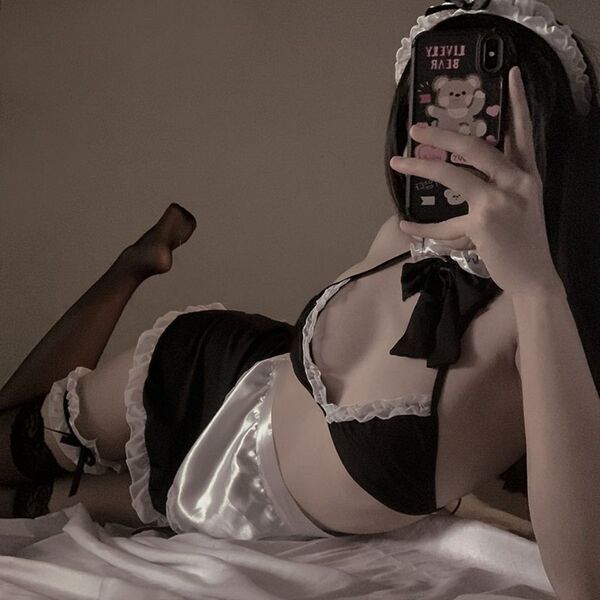 Woman in black and white maid costume with halter neck bow, lace trim, and satin apron, lying on bed taking mirror selfie.
