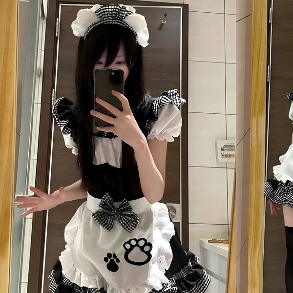 A person wearing a black and white maid costume with ruffles, bows, and paw prints, taking a mirror selfie.