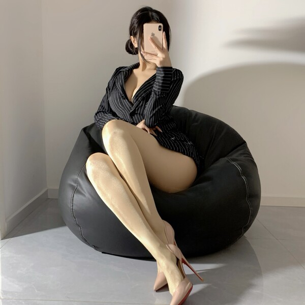 Woman wearing shiny black fishnet stockings with oil-sheen finish, seated on bean bag chair in striped dress and heels.