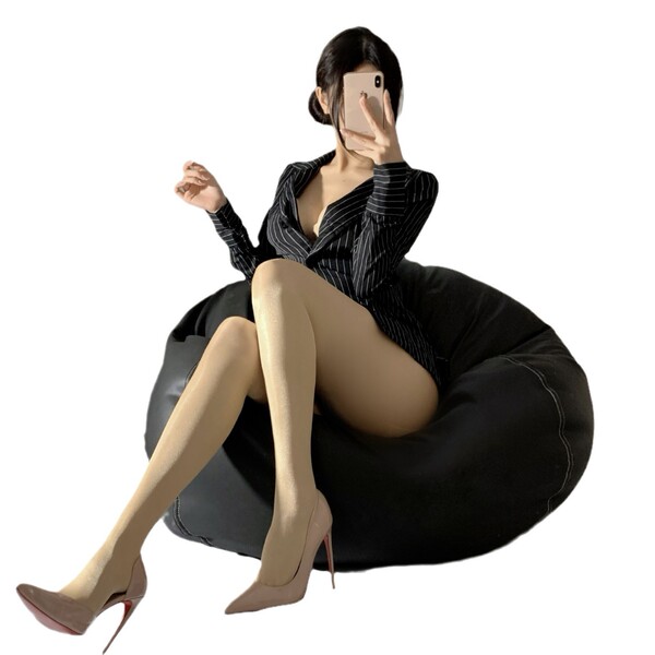 Woman wearing shiny black fishnet stockings with tear-away crotch, paired with a striped dress and heels, sitting on a bean b...