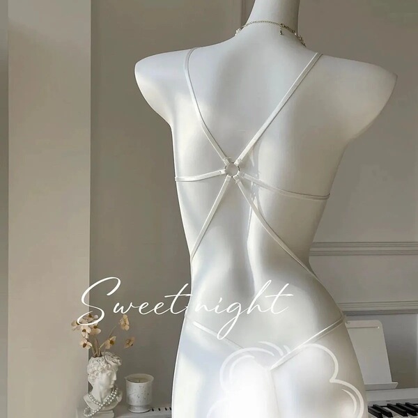White sheer lace bodysuit with crisscross back straps, displayed on a mannequin in a soft-lit bedroom setting.