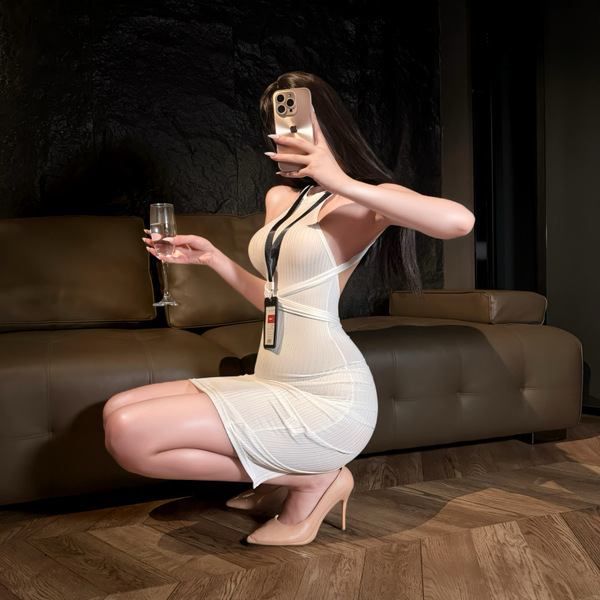 Woman in white summer yang seductive lingerie, holding wine glass and phone, posing on floor beside sofa.