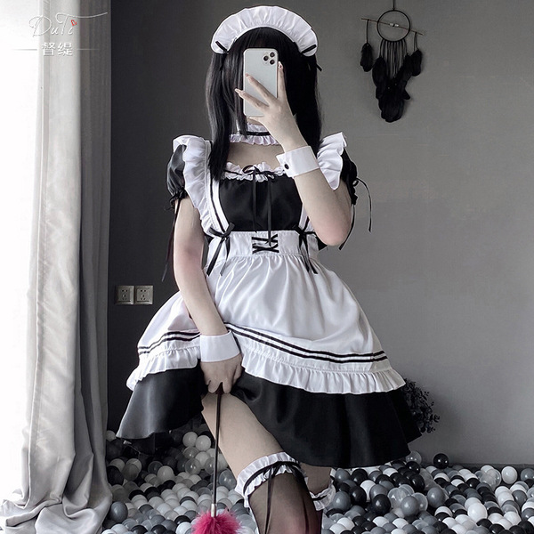 A person wearing a black and white maid costume with ruffled sleeves, apron, and headband, posing indoors near a ball pit.