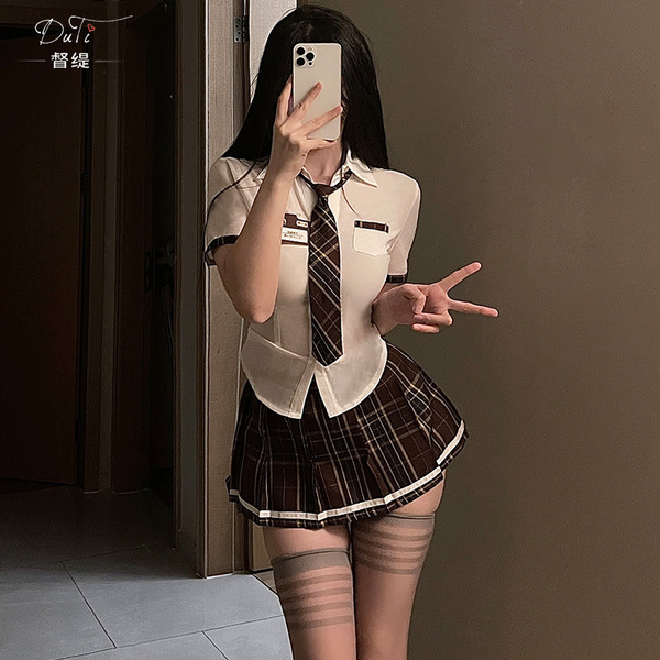 A woman poses with a smartphone, wearing a white blouse, plaid skirt, tie, and stockings, embodying a seductive student unifo...