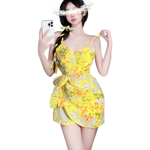 A woman poses with a phone, wearing a yellow floral lace bodysuit set. the outfit features a plunging neckline and a tied wai...