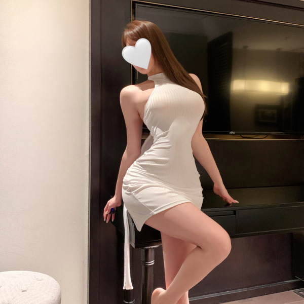 Woman wearing a white, form-fitting sensual lingerie dress with halter neck and side slit, posing in a modern room setting.
