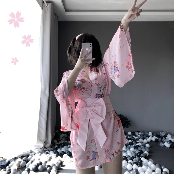 A woman poses with a phone, wearing a pink floral japanese-style kimono robe set featuring lace details and a bow belt. the e...