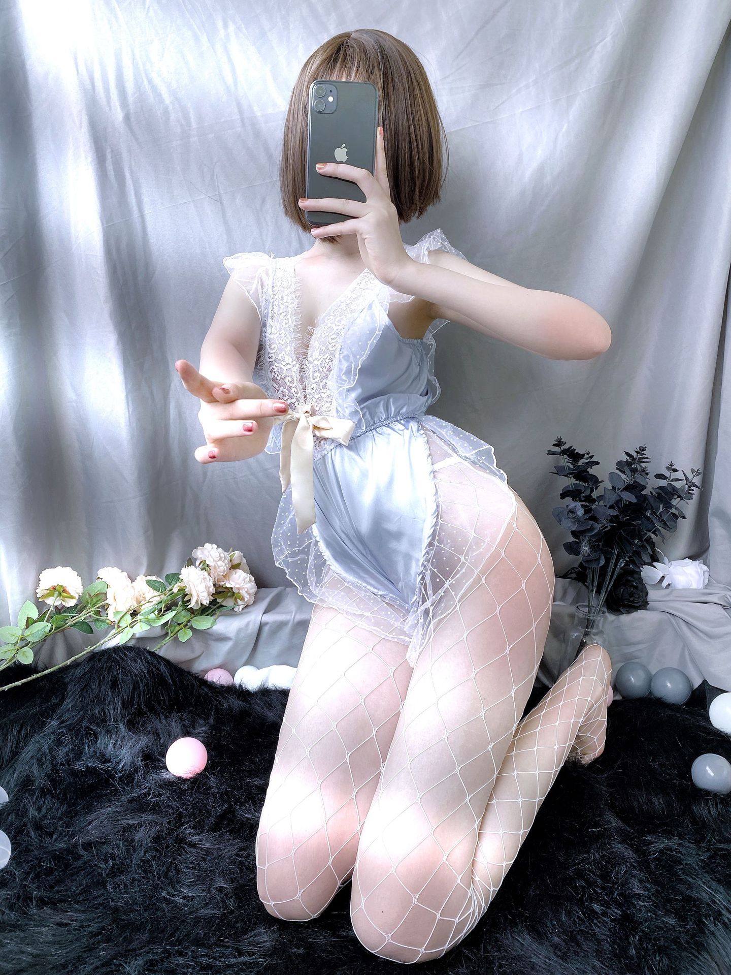 A woman poses with a smartphone, wearing a delicate lace lingerie set paired with fishnet stockings. the outfit features a so...