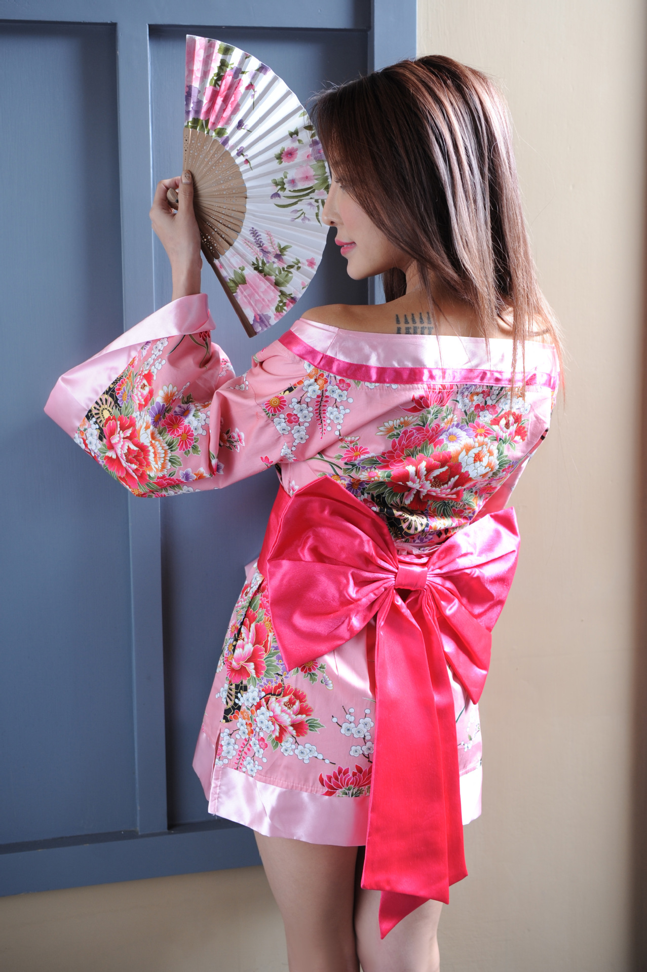 A woman wears a pink floral japanese kimono dress with a large red bow, holding a decorative fan. the outfit features elegant...