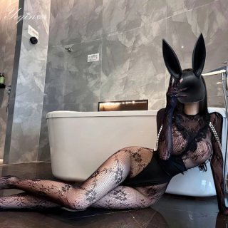 Woman in black lace corset-style mini dress and sheer tights with bunny mask, posing elegantly beside bathtub.