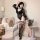 Model wearing black sheer lace bodysuit with garter stockings and holding fan in cozy bedroom setting.