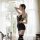 A woman stands by a window holding flowers, wearing a black lace lingerie set with sheer stockings and matching panties, desi...