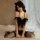 Woman wearing black catwoman lingerie set with feathered top, bow detail, and thigh-high stockings on bed.