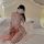 Woman in pink sheer chiffon doudou bralette and matching skirt, sitting on bed with white pillows.