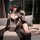 Woman in black fringe lingerie set with sheer sleeves, gold chain belt, and red rose hair accessory on sofa.